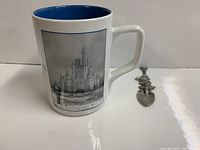 Photo showing the side of the white ceramic mug featuring a grayscale image of Cinderella Castle and the collectible spoon beside it.