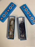 Two blue rectangular boxes with snowflake designs holding two necklaces, one multicolored beaded and one blue beaded.