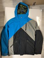 Front view of a Quiksilver snowboarding jacket in blue, gray, and black colors with hood and zippered front pockets, laid flat on a white surface.