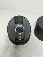Men's wristwatch with stainless steel bracelet and blue dial displayed on a black cushion inside a cylindrical Citizen branded box.