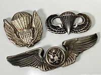 Front view of three silver military insignias: parachute with shield, parachute with wings, and wings with eagle emblem.