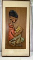 Full view of framed relief wood sculpture showing boy sitting and reading a book, mounted on wood plank.