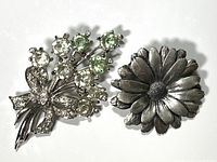 Front view of two silver tone flower brooches, one floral bouquet with rhinestones, the other a solid daisy shape.