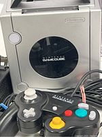 Platinum Nintendo GameCube console with a USB style wired Nintendo controller placed in front, showing the disc lid and controller buttons.