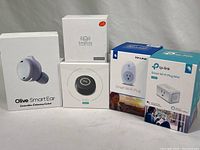 Lot showing all five boxed items: Olive Smart Ear Earbud, Eufy Genie speaker, Simplicity speaker, two different TP-Link smart plugs