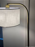 Close-up of the top part of the floor lamp showing the fabric shade with LED bulb inside, mounted on a curved arm with a gold finish.