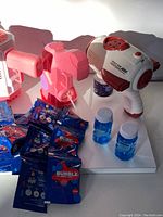 Photo showing three bubble guns in white, pink, and light pink colors along with multiple blue bubble solution trading card packs and two small bottles of blue bubble solution liquid.