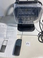 LED search light with protective clear plastic cover, remote control, and instruction manual placed on a flat surface indoors.