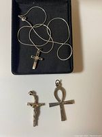 Top view of silver chain necklace with cross pendant lying in black box, with two additional cross-shaped pendants beside it.