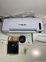 White TOPELEK thermal laminator with accessories including wooden clips, small metal pins, black pouch, funnel-shaped tool, and user manual assembled on a white surface.