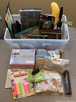 Overview of various miscellaneous household items placed inside a transparent plastic box with no lid.