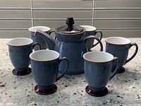 Front view of the Denby teapot surrounded by six matching teacups on a granite countertop with tiled backsplash.