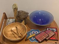 View of all items in lot showing pelican ceramic figure, olive wood bowl with tongs, blue glass bowl, Paris-themed ceramic catch-all, grapefruit spoons, cocktail forks, sharpener, and trivet.