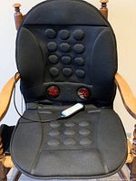 Full view of black massage cushion on wooden chair showing rounded massage nodes on seat and back with two red circular elements at lumbar area and attached remote.