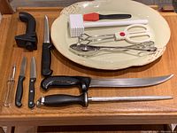 Overview of the lot showing Casafina oval platter, multiple cutting and carving knives with black handles, sharpening steel, meat tenderizer, kitchen scissors with white handles, metal utensils with white and red handles.