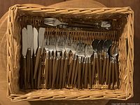 Top-down photo of the full cutlery set laid inside the wicker basket showing knives, forks, teaspoons, and some serving utensils all with brown composite/plastic handles.