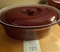 Cranberry-colored ceramic casserole dish with lid, closed, on wooden surface with auction tag number 121 visible.
