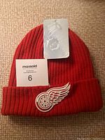 Front view of the red toque with NHL tag, folded cuff displaying embroidered Detroit Red Wings winged wheel logo in white and silver thread.