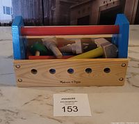 Front view of Melissa & Doug wooden toolbox filled with wooden toy tools, showing blue sides, red handle, and natural wood front branded panel.