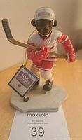Front view of the bear figurine in Detroit Red Wings uniform, holding hockey stick on rink base with tag visible.