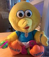 Front view of Baby Big Bird plush showing yellow face, blue shirt, and colorful rattle toy in hand. Text on feet visible: 'crinkle' on left foot, 'rattle' on right foot.