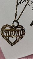 Close-up of the gold-tone heart pendant featuring cut-out "MOM" text and smaller heart inside. Shows detail and condition of pendant and part of chain.