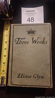 Front cover of the book showing title 'Three Weeks' by Elinor Glyn with a crown symbol, beige cloth hardcover with black text.