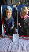 Front view of Bill and Hillary Clinton figures in their original boxes, showing the figures standing upright with Bill Clinton in a dark suit and red tie, Hillary Clinton in a black jacket and pink blouse.