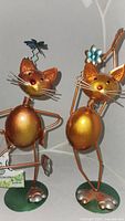 Two copper finish whimsical yoga cat metal figurines with wire whiskers on a green base with decorative head accessories
