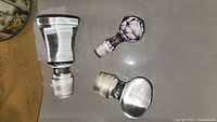 Top view of three assorted bottle stoppers on a surface showing purple cut glass, clear cut glass, and clear crystal stopper.