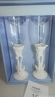 Pair of champagne flutes displayed in a box, showing clear glass cup and white swan detailed bases forming an embracing pose.