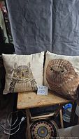 Two 16x16 inch throw pillows with embroidered owl faces seated on a wooden stool against a gray backdrop