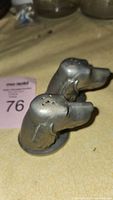 View of two pewter dog head salt and pepper shakers from the front on a yellow textured surface with auction tag visible.