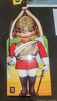 Photo of a vintage postcard showing a cartoon soldier in red uniform with gold helmet and white sash, holding a sword