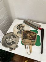 Photo showing four antique hand fans arranged on a white surface. 2 circular portrait hand fans, 1 rectangular biblical art fan, 1 folded fabric fan with red tassel.