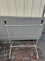 Full view of white metal standing towel rack showing two horizontal bars for hanging towels and four curved feet for stability, placed on brick flooring in outdoor lighting.