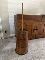 Full view of antique wooden butter churner standing on carpeted floor, showing the tapered barrel and long plunger.