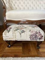 Front view of floral upholstered footstool with carved wooden legs in front of sofa.