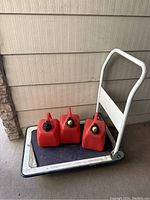 Photo of 3 red plastic gas canisters and one white hand truck dolly on wooden floor near wall