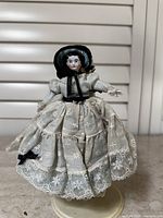 Front view of antique china head doll showing white embroidered dress with lace and black bonnet with ribbon ties.