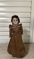 Front view of the bisque doll wearing brown dress and original wig standing on display stand.