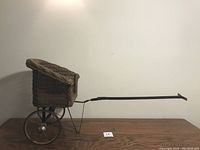 Side view of the wicker doll carriage with large metal wheels and a long handle with a wicker cover partially folded back.