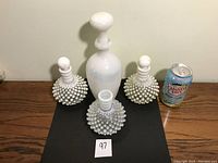 Four antique milk glass bottles: two hobnail globe-shaped with stoppers, one hobnail globe-shaped without stopper, and one tall smooth milk glass bottle with stopper. Canada Dry soda can included for scale.