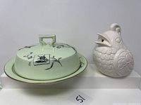 Front view of covered cheese dish with plate showing black illustration design and bird-shaped ceramic dispenser beside it.