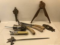 View of the nutcracker/clam opener tool with wooden handles, yellow pocket knife, bird-handle letter opener, and other assorted metal vintage items on white surface.