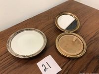 Two circular compacts on wooden surface: silver-tone embossed compact (closed) and gold-tone green enamel compact opened showing mirror and powder tray.
