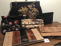 Photo showing seven vintage bags and clutches arranged on a wooden surface. Includes the Simpson's France beaded clutch with floral and leaf design, black floral embroidered clutch, snakeskin pattern clutch, black fabric envelope clutch with gold letter 'B', and multiple leather clutches including crocodile embossed style.
