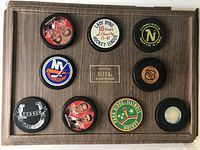 Two wooden holders each with 9 hockey pucks featuring various logos and designs including NHL teams and other hockey organizations.