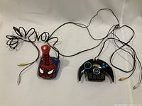 Two Jakks Pacific plug-and-play game controllers laid flat, Spider-Man joystick in red and blue, Batman controller in black with blue buttons, all cables connected.