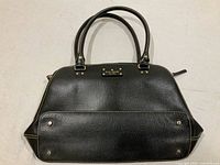 Photo of the handbag exterior showing black pebbled leather, dual rolled handles, gold-tone hardware, and base with brass feet.